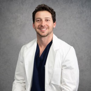 Studio headshot of Dr. Chaz Horton smiling in a white lab coat over scrubs against a grey backdrop