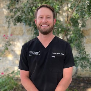 Portrait of Dr. Seth Harris smiling outdoors in dark scrubs against a stone wall and green leaves background